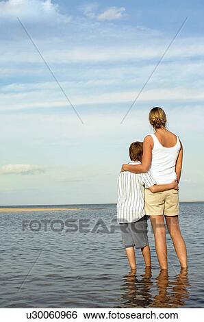 Mother and Son standing in water by shore of Beach looking at view, back view View Large Photo Image Stock Photograph - Mother and Son standing in water by shore of Beach looking at view, back view. Fotosearch