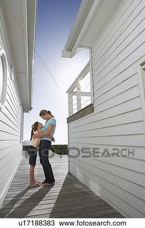 Stock Image - Mother embracing daughter in passageway between houses, side view. Fotosearch
