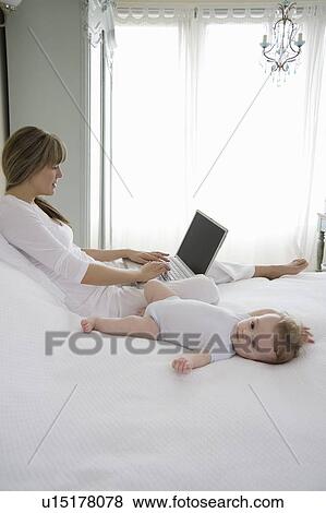 Stock Photo - Mother sits typing on laptop with baby beside her on bed. Fotosearch