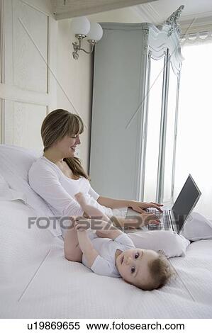 Stock Photography - Mother sits typing on laptop with baby beside her on bed. Fotosearch