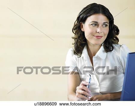 Stock Photo - Office worker holding phone and using laptop. Fotosearch