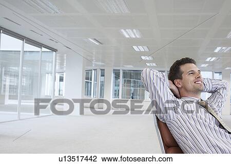 Stock Image - Office worker leaning back in swivel chair, hands behind head, in empty office space. Fotosearch