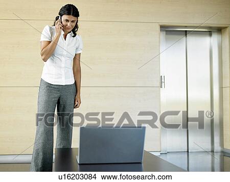 Picture - Office worker standing in lobby by laptop using cell phone. Fotosearch