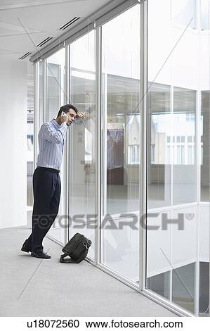 Stock Image - Office worker talking on mobile phone, looking out window of empty office space. Fotosearch