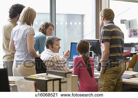 Stock Image - Office workers standing around colleague using computer, back view. Fotosearch