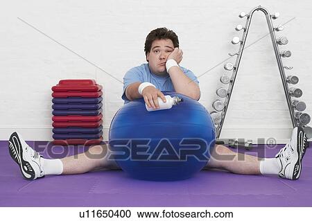 Stock Image - Overweight Man sitting on floor with exercise ball in health club, portrait. Fotosearch