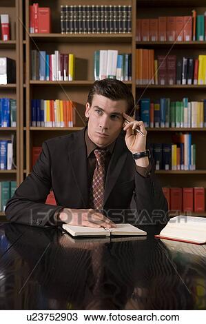 Stock Image - Pensive man wearing suit with book at desk in library. Fotosearch