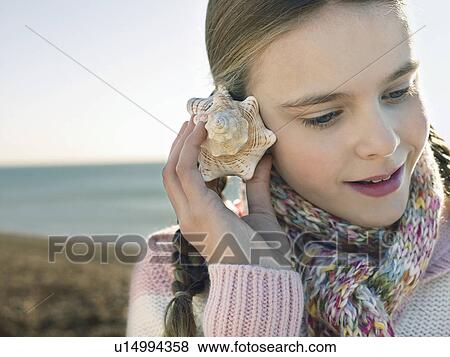 Stock Photo - Pre teen girl listening to seashell, standing on beach, close up. Fotosearch