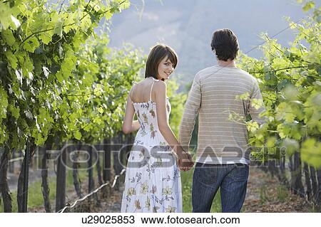 Stock Image - Rear view of young couple walking through vineyard hand in hand, woman smiling back over her shoulder. Fotosearch