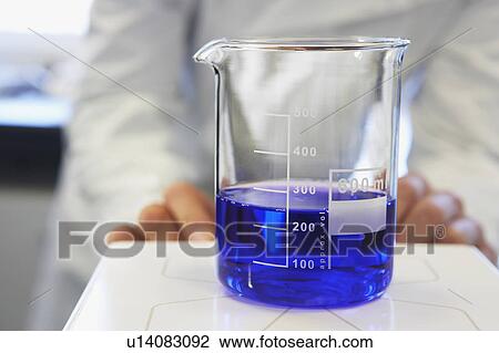 Stock Image - Scientific glass container with blue liquid, technician in background. Fotosearch