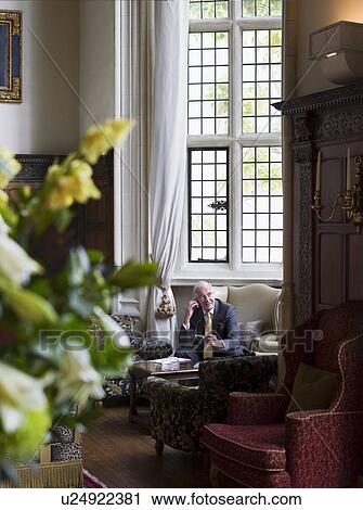 Stock Image - Senior man sitting in interior with antique furniture, using mobile phone. Fotosearch