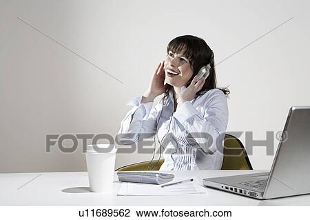 Smiling woman with earphones sitting at desk View Large Photo Image Stock Image - Smiling woman with earphones sitting at desk. Fotosearch