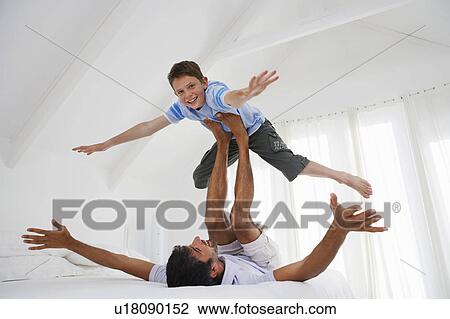 Stock Image - Son balancing on father's legs on bed in bedroom. Fotosearch