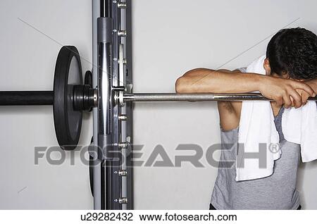 Stock Image - Sweaty man resting on barbell after workout. Fotosearch
