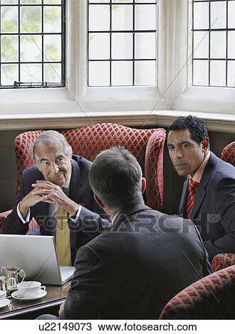 Stock Image - Three business men talking over laptop in lobby. Fotosearch