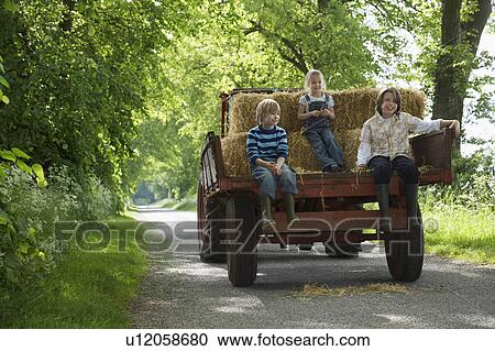 Stock Image - Three children (5-9) sitting on trailer on country lane. Fotosearch
