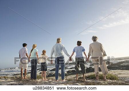Stock Image - Three generation family holding hands, on seashore, back view. Fotosearch
