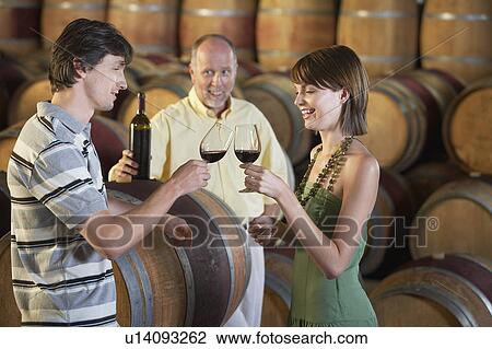 Stock Image - Three people wine-tasting beside wine casks. Fotosearch