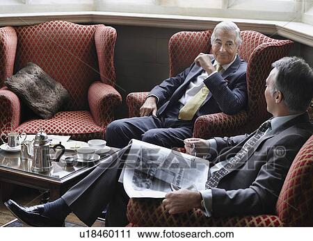 Stock Image - Two business men sitting in lobby, talking, one holding newspaper. Fotosearch