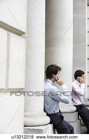 Stock Photo - Two businessmen on cell phones sitting on pillars outside building. Fotosearch