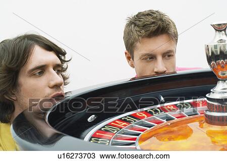 Stock Image - Two hopeful young men crouching at roulette wheel watching spin, close up. Fotosearch