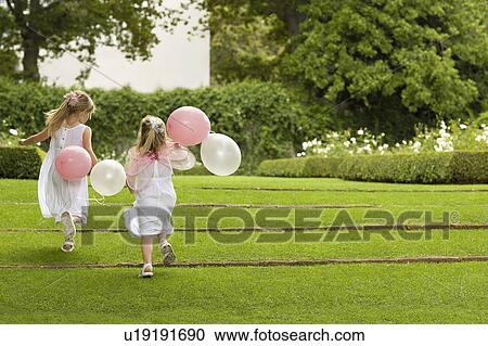 Stock Image - Two young girls running in garden, holding balloons, back view. Fotosearch