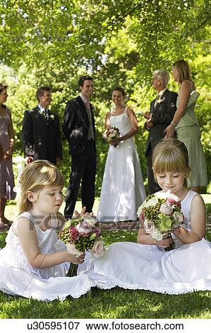 Stock Photo - Two young girls sitting on lawn, holding bouquets, bride and groom in background. Fotosearch