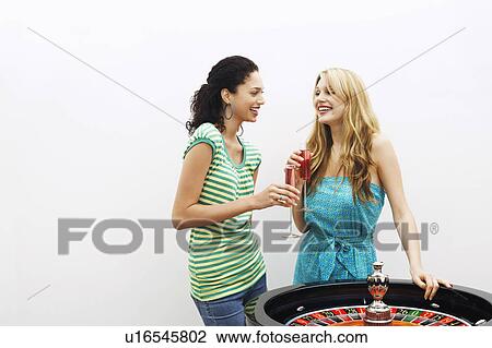 Stock Image - Two young women drinking cocktails by roulette wheel. Fotosearch
