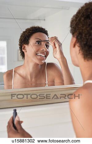 Stock Image - Woman applying mascara in mirror, at home. Fotosearch