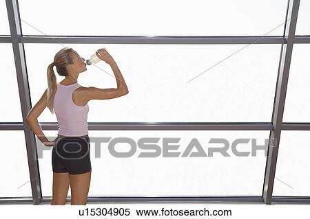 Stock Photography - Woman drinking from water bottle by window in health club. Fotosearch