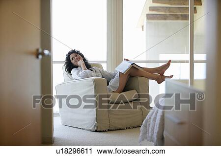 Stock Image - Woman holding book, sitting on armchair by window, indoors. Fotosearch