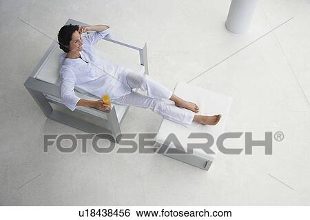 Stock Photograph - Woman holding glass of juice, reclining in armchair, elevated view. Fotosearch