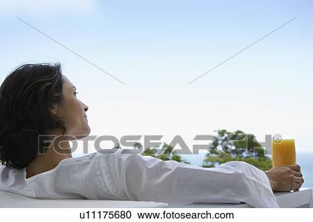 Stock Image - Woman holding glass of orange juice, sitting outdoors, back view. Fotosearch