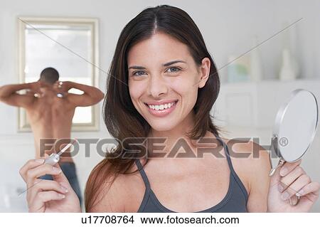 Picture - Woman holding lip gloss and mirror in bathroom, portrait. Fotosearch