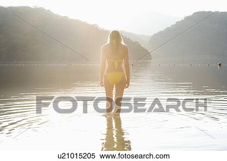 Stock Photography - Woman in yellow bikini stands at edge of lake. Fotosearch