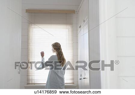 Stock Photo - Woman peering through blinds standing at window, half length. Fotosearch