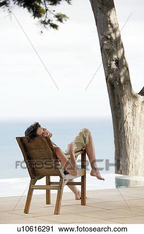 Stock Image - Woman sitting on lounge chair by infinity pool overlooking sea. Fotosearch