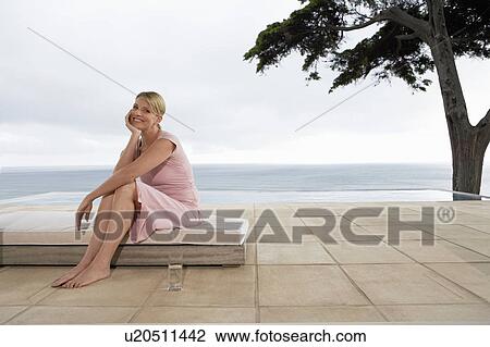 Stock Image - Woman sitting on sun bed by infinity pool, portrait. Fotosearch