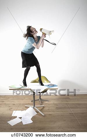 Stock Image - Woman standing on desk, shouting through megaphone. Fotosearch