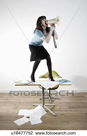 Stock Image - Woman standing on desk, shouting through megaphone. Fotosearch