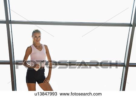 Stock Image - Woman standing with water bottle by window in health club, portrait. Fotosearch