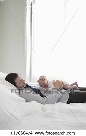 Picture - Working father lies with two week newborn on bed. Fotosearch