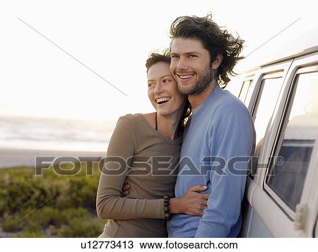 Stock Image - Young couple embracing by van parked by ocean, half length. Fotosearch
