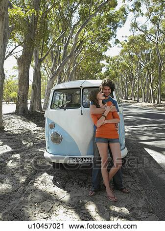 Stock Image - Young couple embracing, leaning against front of camper van parked by road. Fotosearch