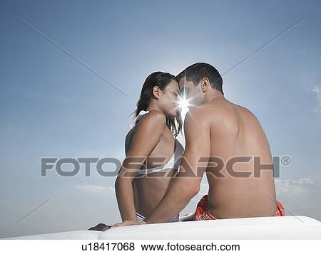 Stock Photo - Young couple embracing on cushions outdoors, face to face, sun breaking through, back view. Fotosearch