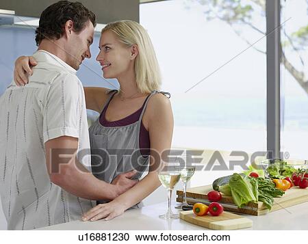 Stock Image - Young couple hugging in kitchen near countertop with fresh produce and wineglasses. Fotosearch