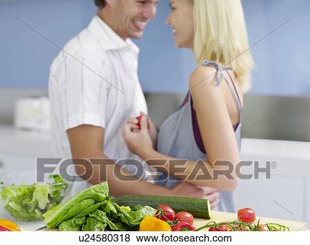 Stock Photo - Young couple standing face to face, Flirting in Kitchen near fresh produce, side view. Fotosearch