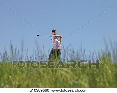 Stock Image - Young female golfer driving ball, low angle view. Fotosearch