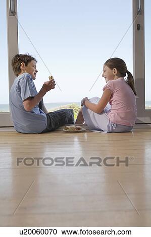 Stock Image - Young girl and boy sitting on floor in doorway eating sandwiches. Fotosearch