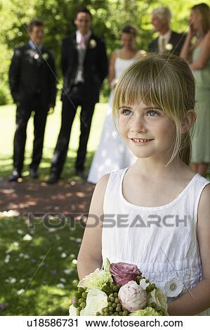 Stock Image - Young girl holding bouquet, bride and groom in background. Fotosearch